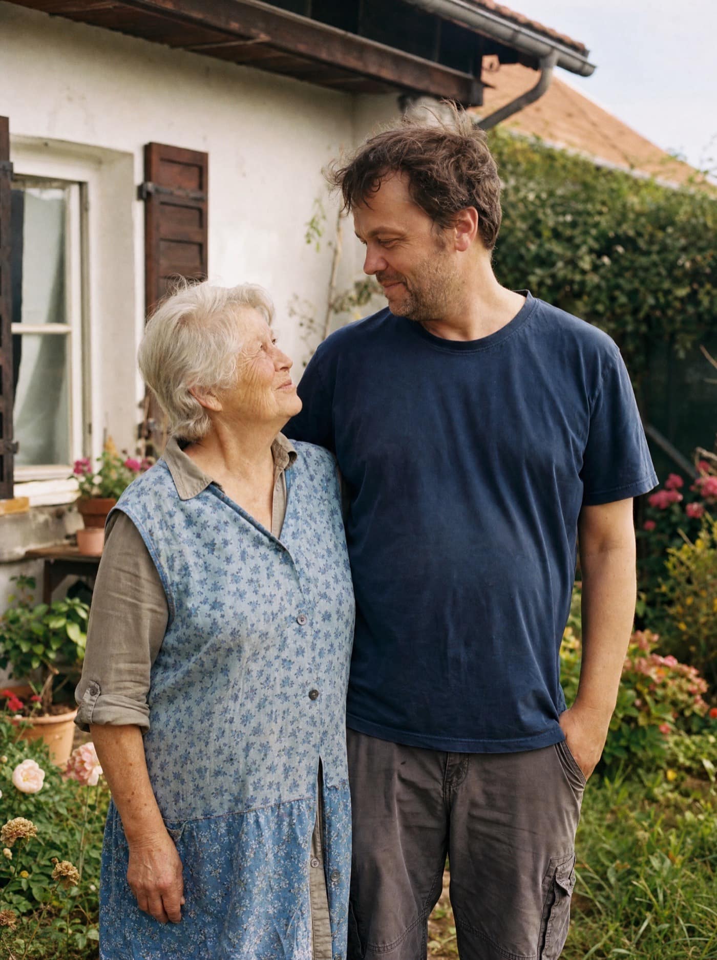 Eine ältere Frau und ihr erwachsener Sohn vor einem Quierschieder Wohnhaus, im warmen Licht des Nachmittags. Verbundenheit über Generationen, ruhig und echt.